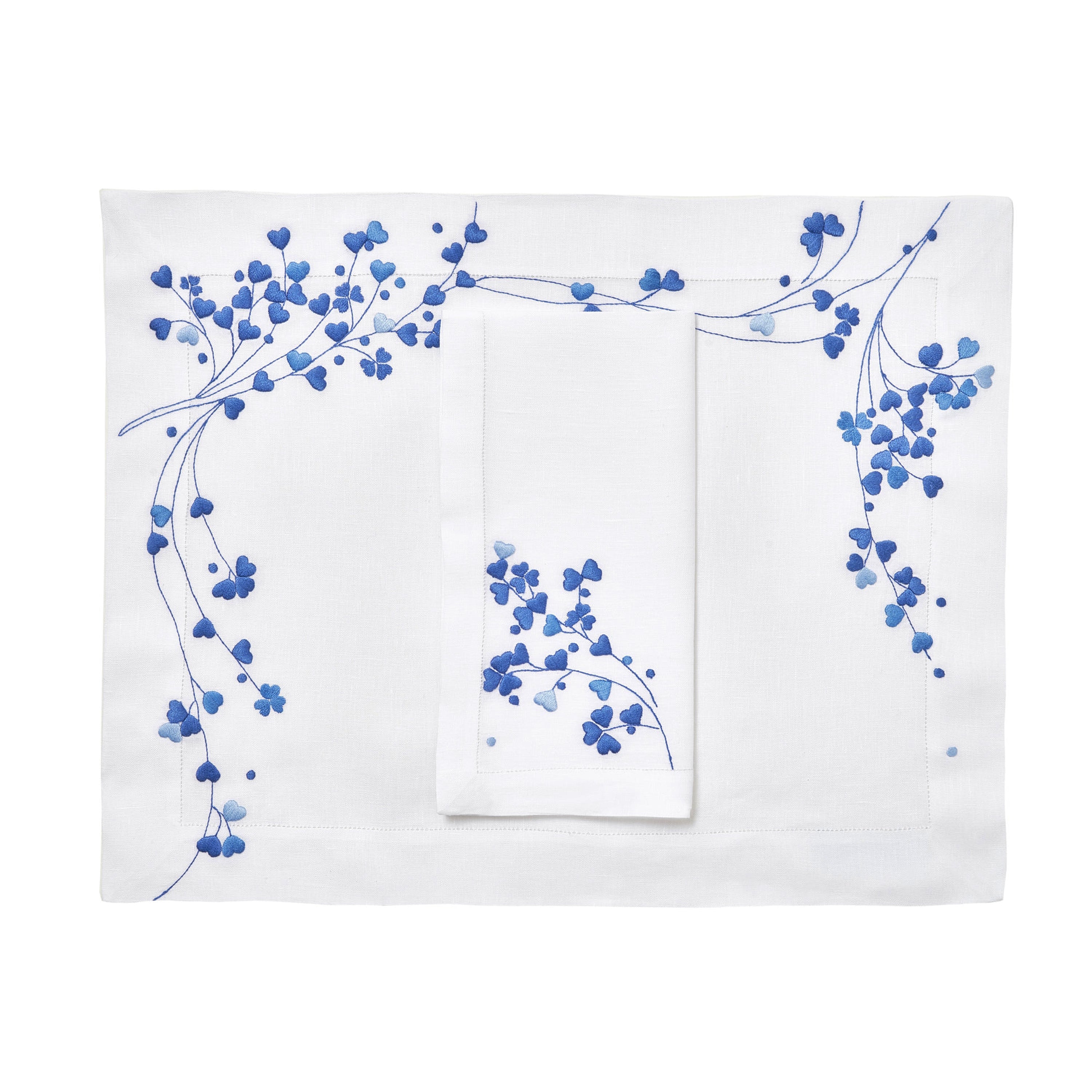 Blue embroidered hearts, Anagramme Embroidery, shown on placemat with a folded napkin, finished with a hemstitch on white linen, on a white background.