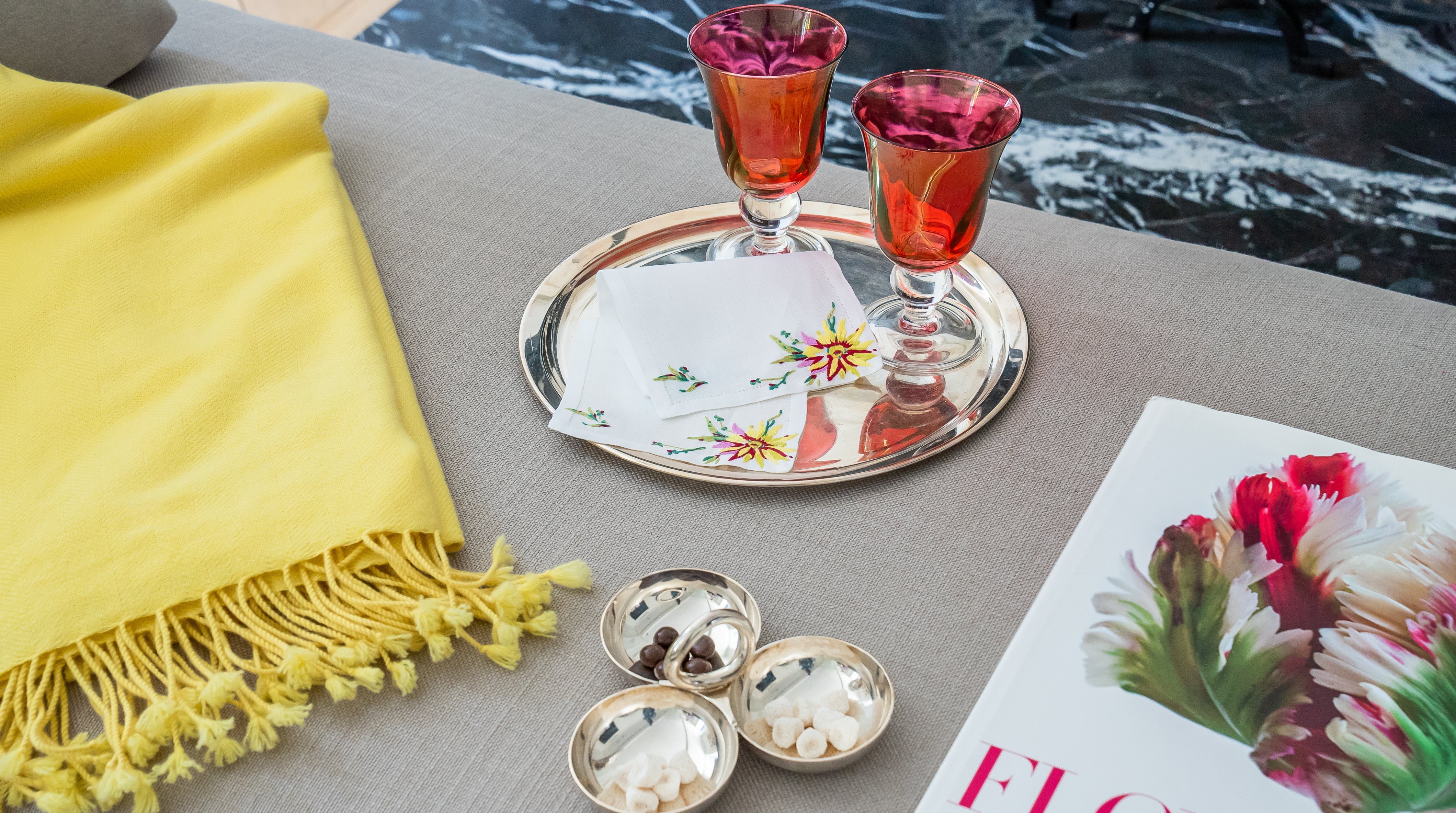 Decorative setting with red glasses, silver tray, yellow blanket, and floral book on a gray surface.