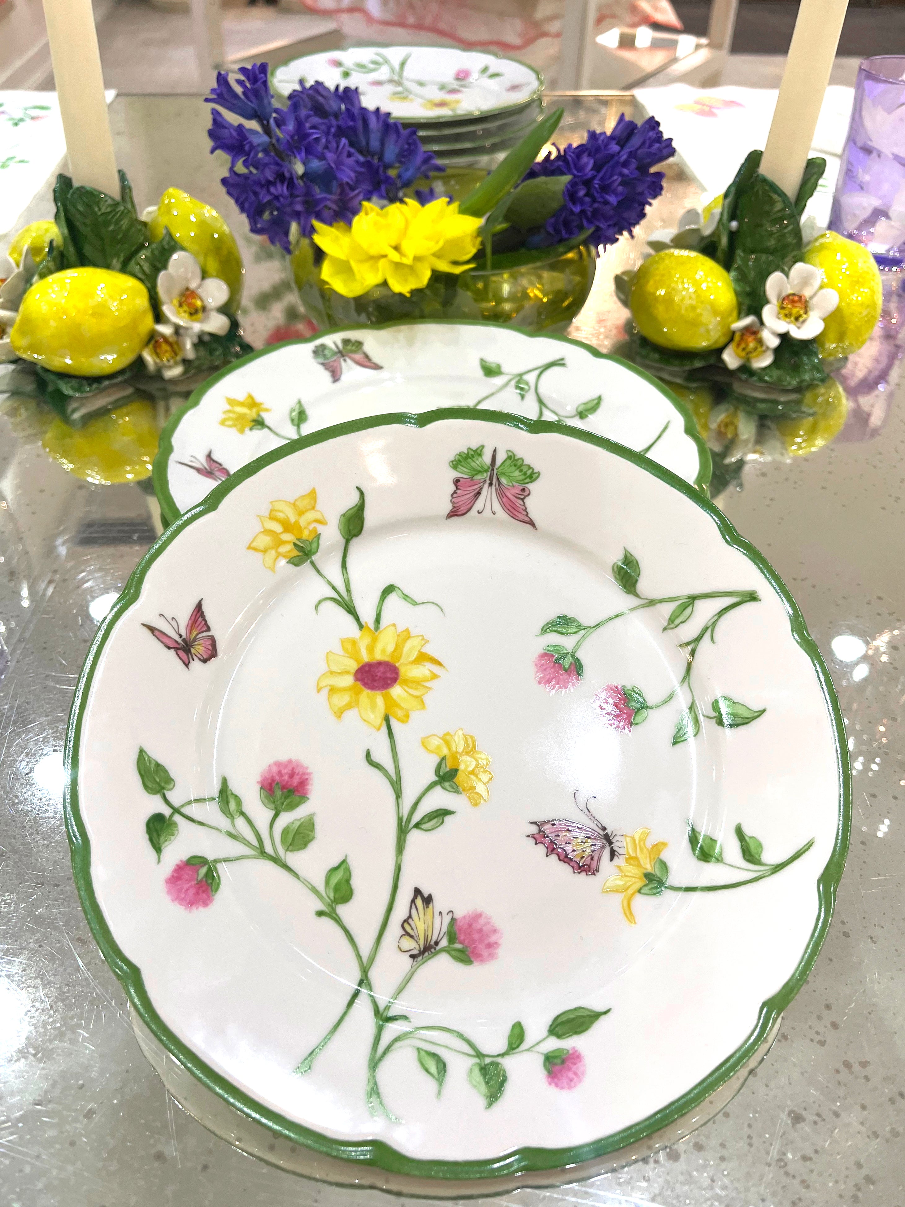 Printed florals and butterflies (in pink, yellow, lilac and green) on porcelain dessert plates, on glass table with yellow and purple florals. lemon candlestick holders with white stem candle.