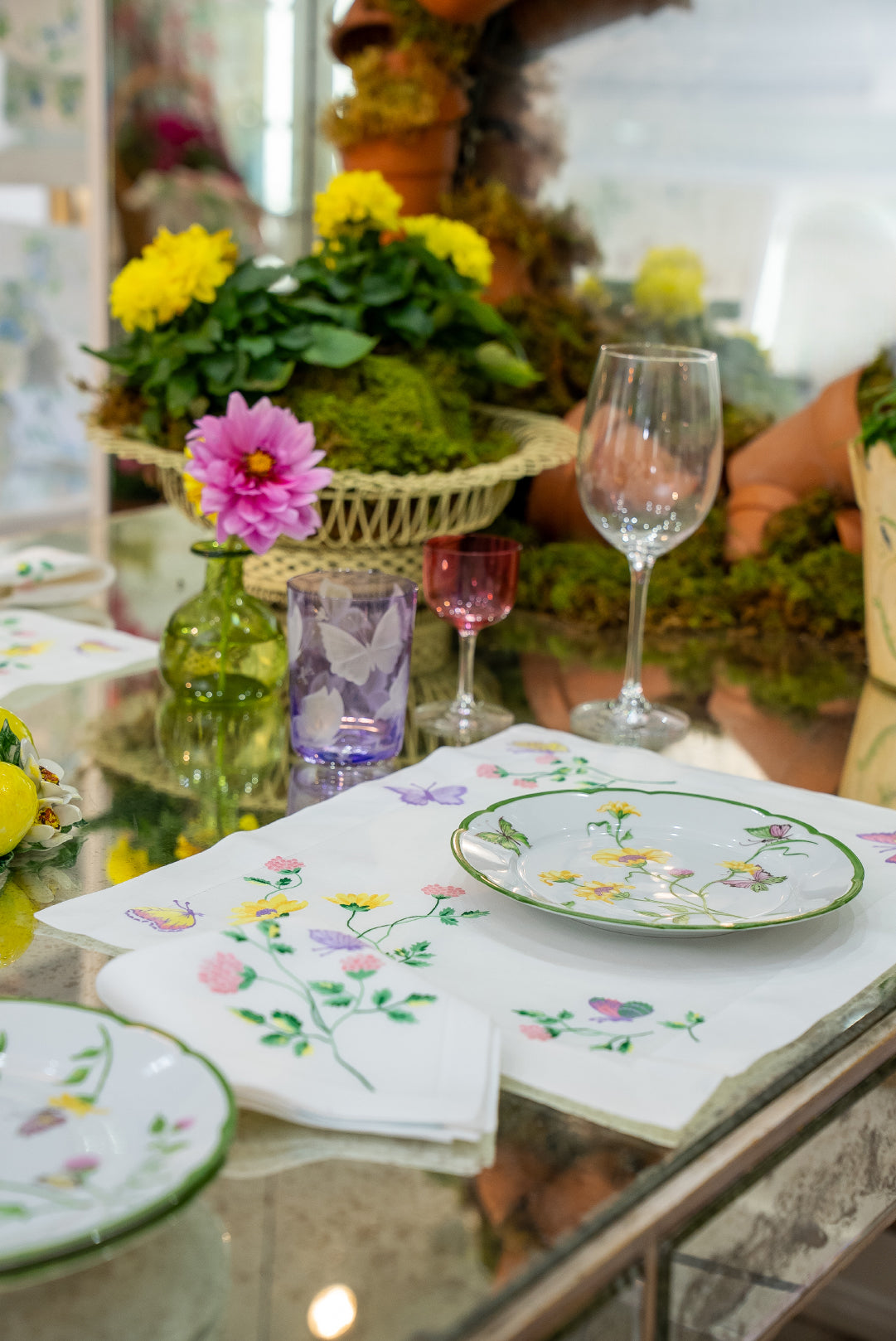 Embroidered florals and butterflies (in pink, yellow, lilac and green) Table linens displayed on glass table, with fresh flowers, and assorted colored glass stemware.