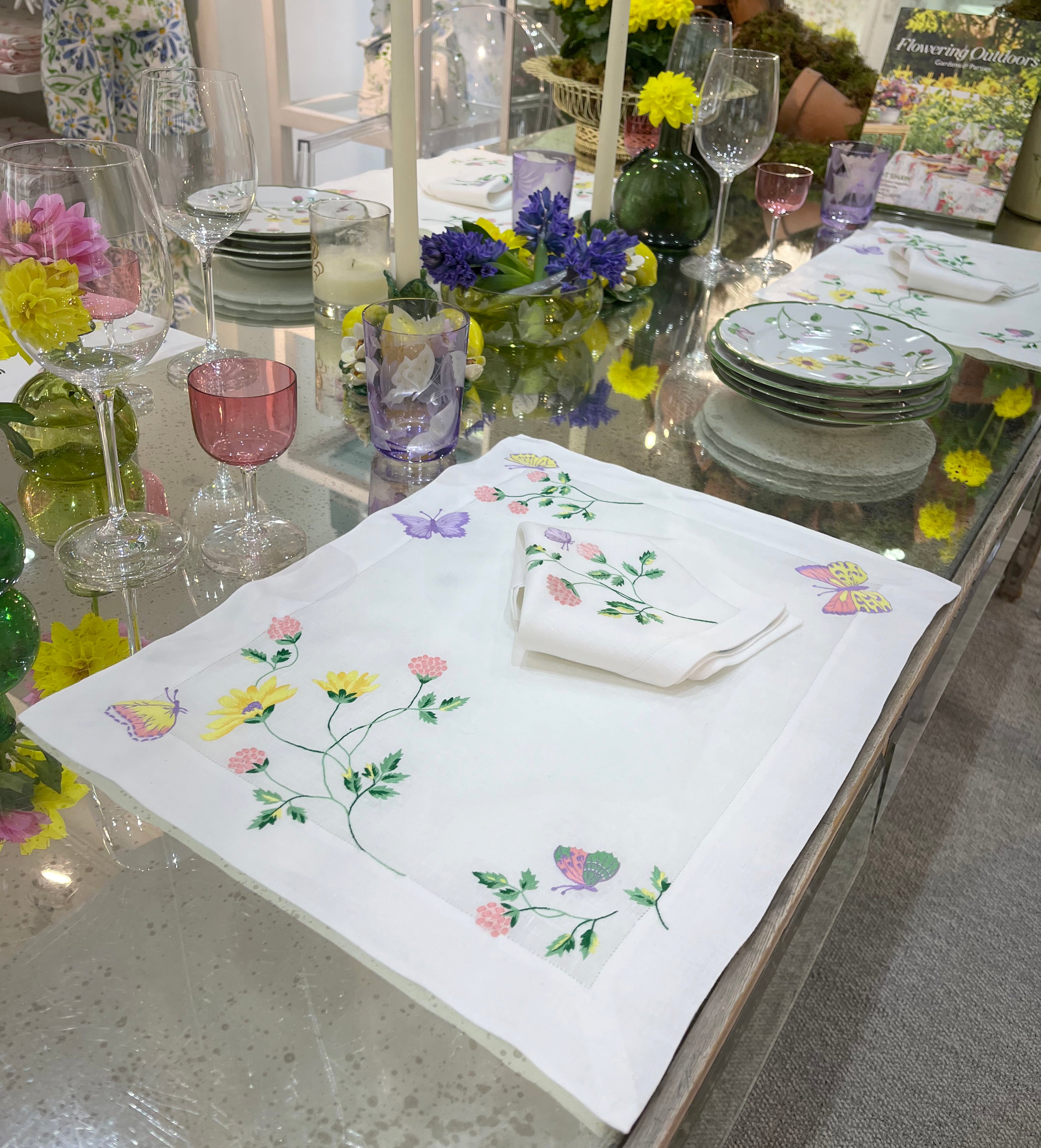 Embroidered florals and butterflies (in pink, yellow, lilac and green) Table linens displayed on glass table, with fresh flowers, and assorted colored glass stemware. Flowering Outdoor book by Margot Sham in background.