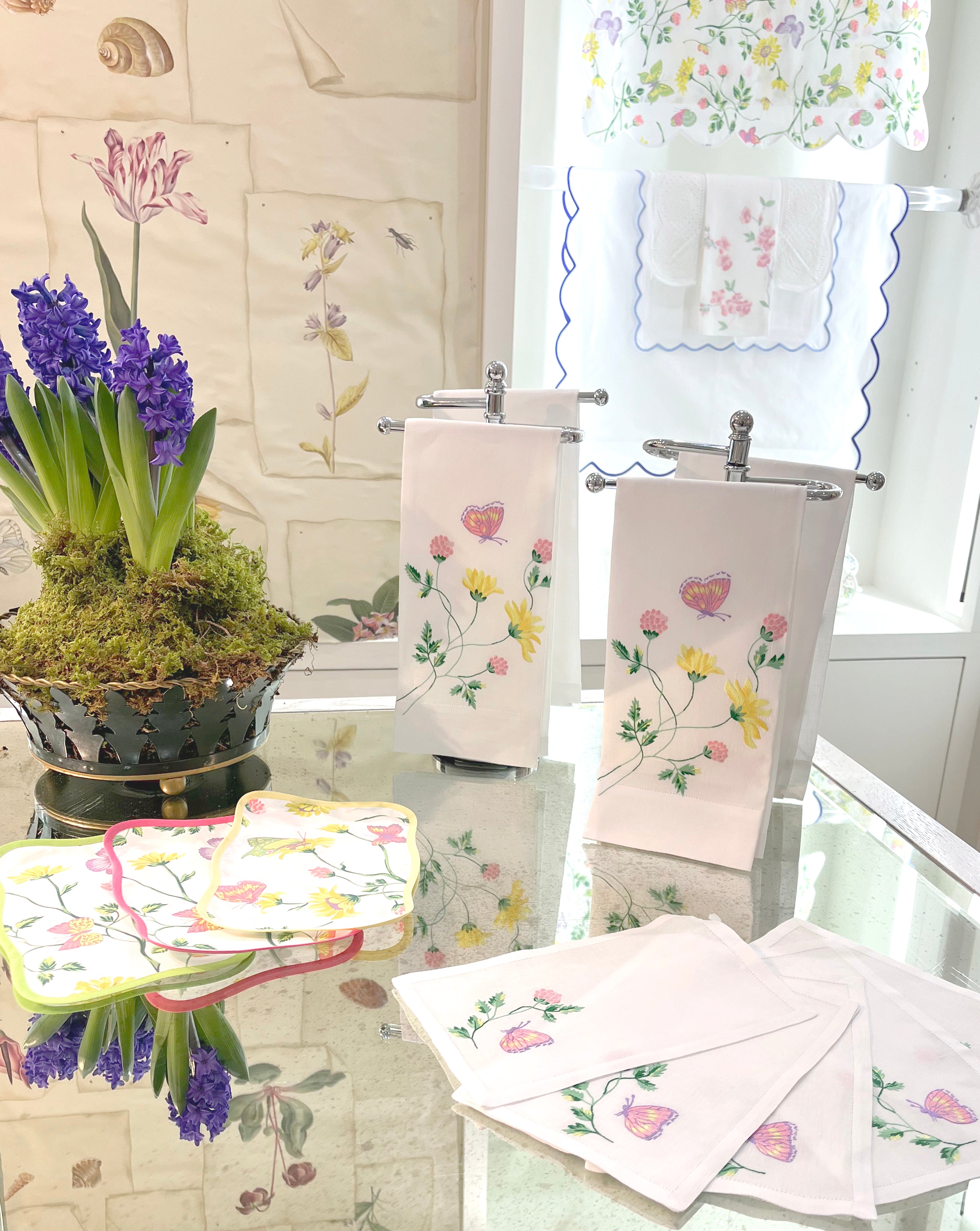 Printed and embroidered florals and butterflies (in pink, yellow, lilac and green) table linens displayed on glass table. with coordinating shams on back, and de Gournay Wallpaper (Bunny Mellon Collection).