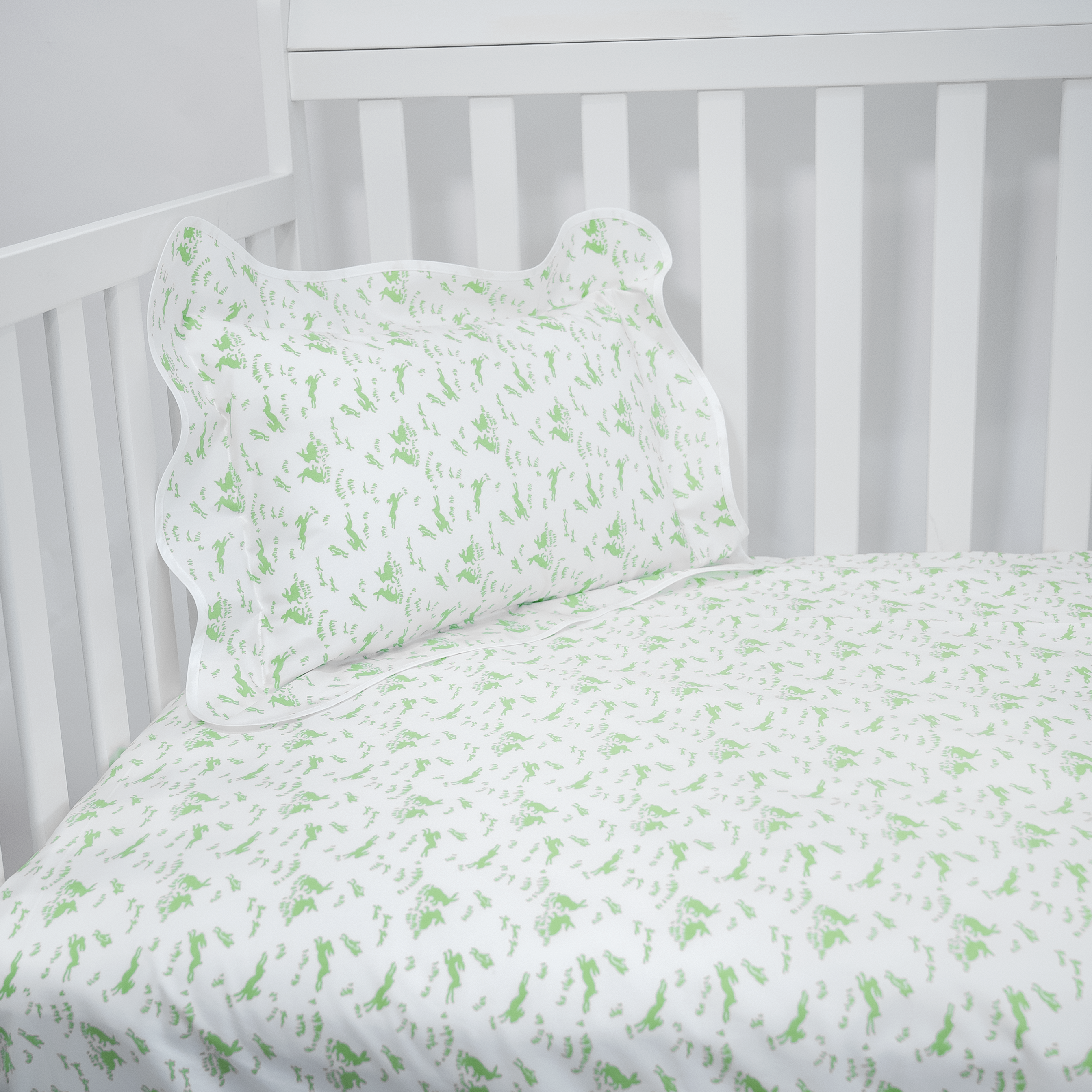 Printed Soft green Lapins print (bunnies) on crib fitted sheet, corner shot of crib with wavy white boudoir in matching print.