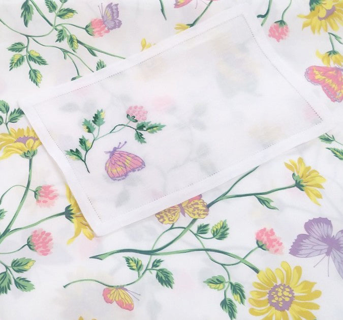 Printed and embroidered florals and butterflies (in pink, yellow, lilac and green) cocktail napkin displayed on fabric.