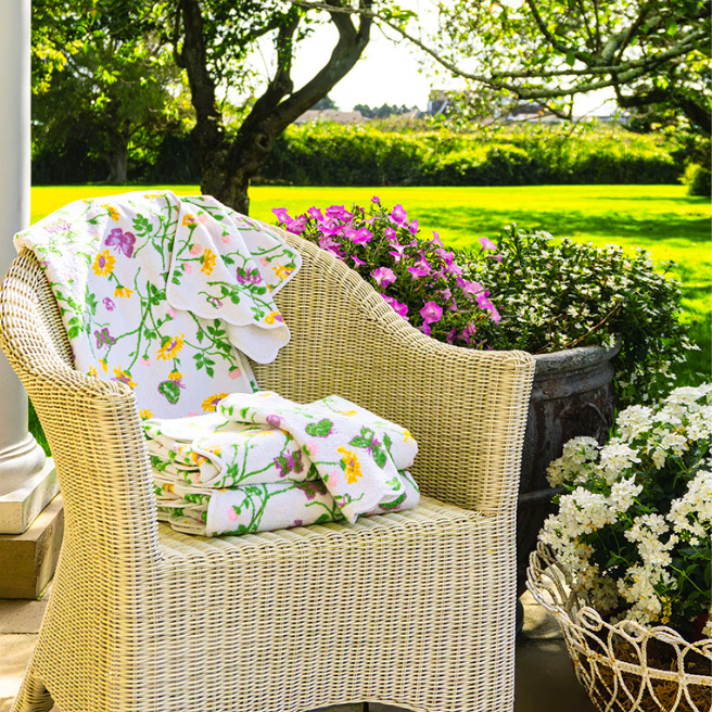 Sunlit wicker armchair draped with a floral quilt sits on a garden patio, surrounded by pink and white flowers in a peaceful setting.