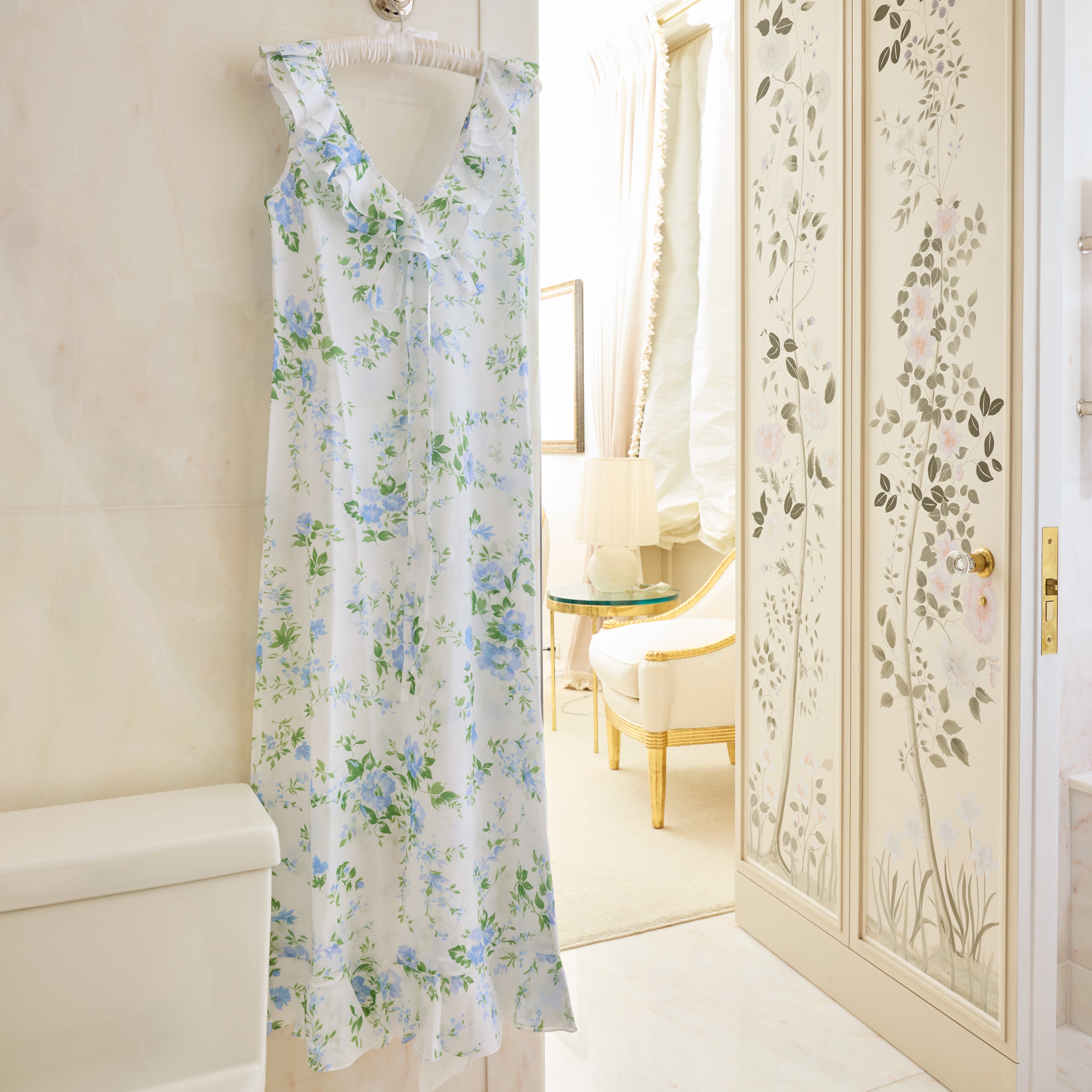 A light blue and green floral dress hangs on a hook in a marble bathroom, with a toilet nearby and decorative painted closet doors in the background.