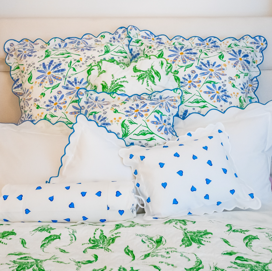 Elegant bed styled with white linens featuring blue and green floral motifs (Marguerites Blue/Green, and Muguet Green) with a scalloped blue trim, paired with Coeurs Blue heart-print pillows and bolsters. Crisp white sheets and embroidered details create a fresh, refined bedroom setting
