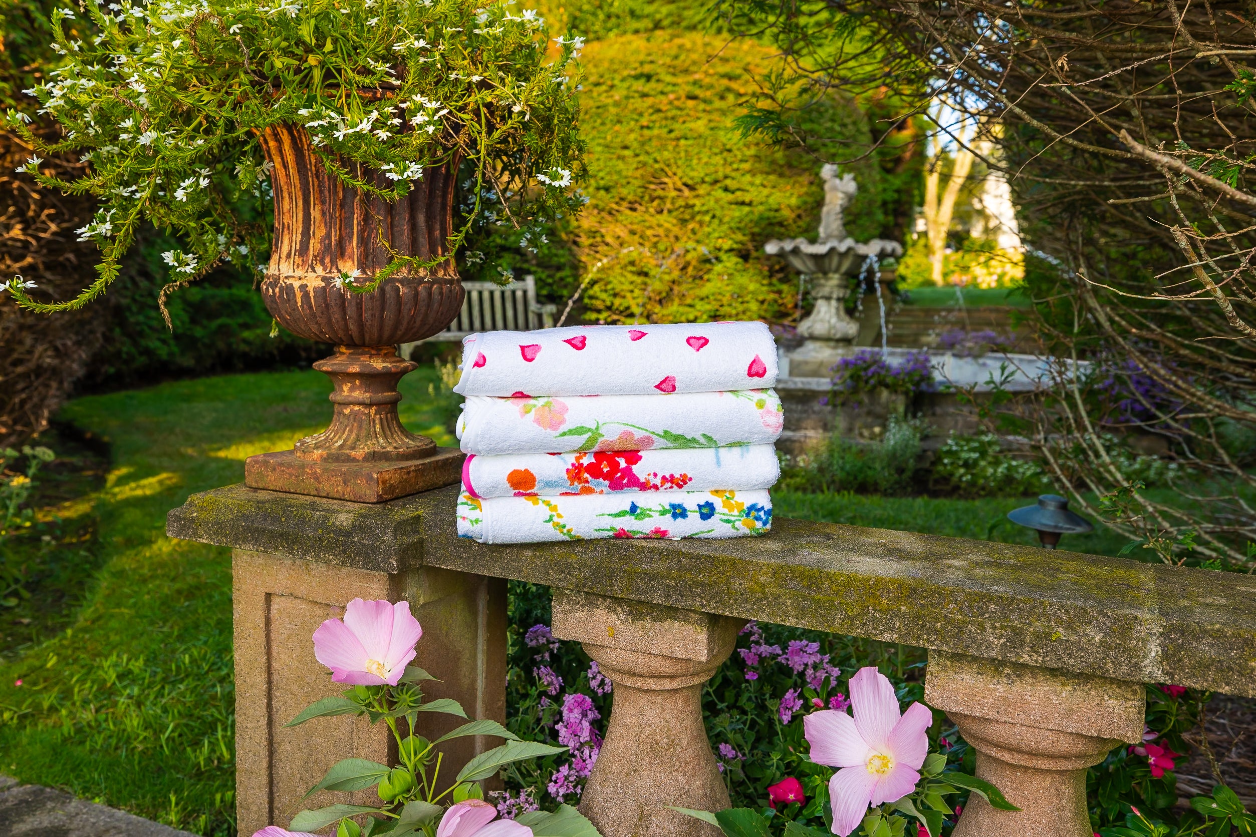 Stack of floral-patterned towels on a stone table in a garden setting with a fountain in the background.