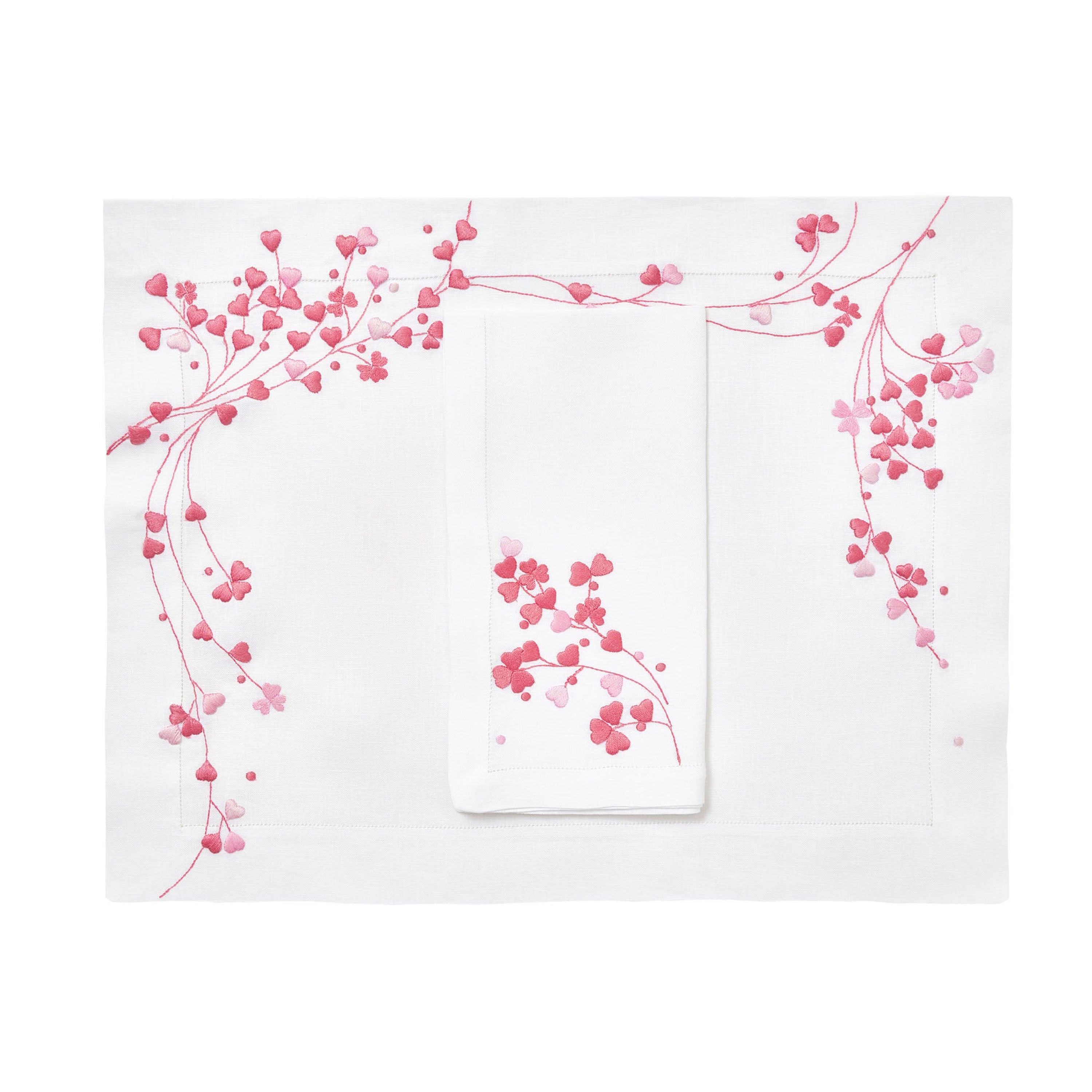 Pink embroidered hearts, Anagramme Embroidery, shown on placemat with a folded napkin, finished with a hemstitch on white linen, on a white background.