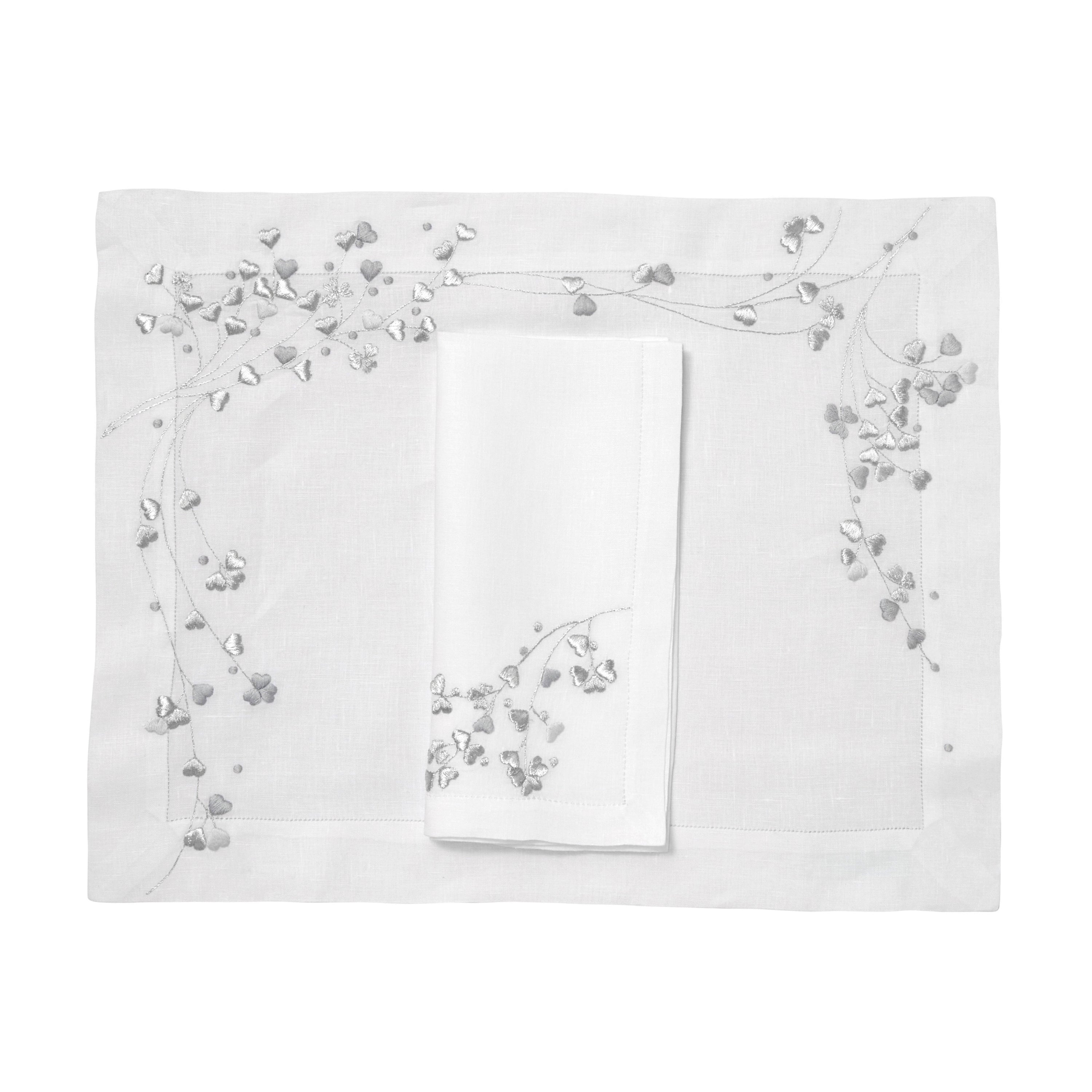 Silver and grey embroidered hearts, Anagramme Embroidery, shown on placemat with a folded napkin, finished with a hemstitch on white linen, on a white background.
