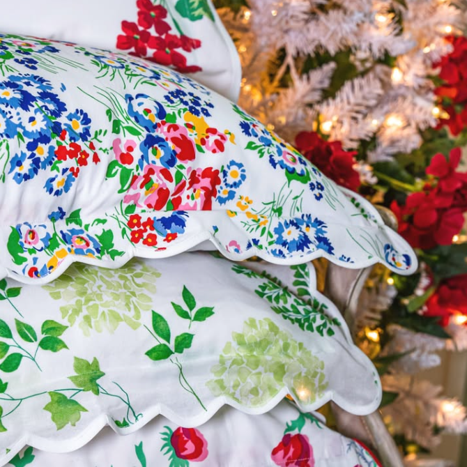 Stacked decorative pillows featuring Geraniums, New York Mille Fleurs Multi, Mariage, and Coquelicots Parisiens prints, finished with signature scalloped edges and styled beside a softly lit holiday tree.