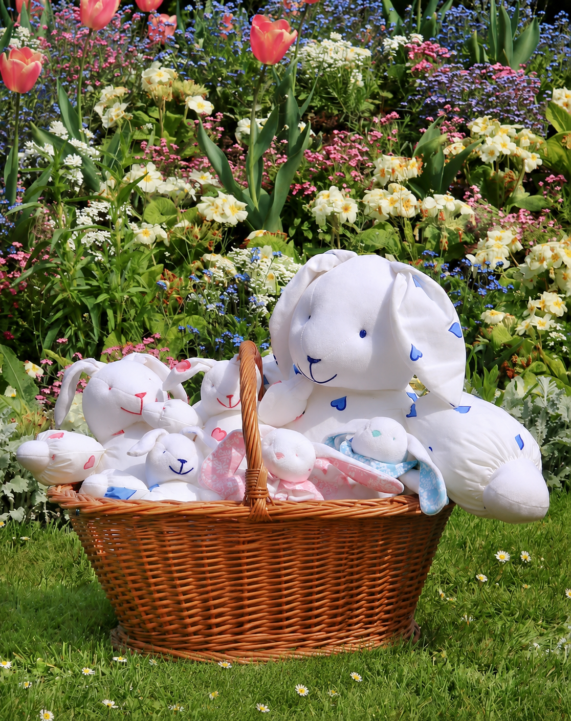 A wicker basket filled with stuffed bunny toys sits on grass in front of a garden with blooming flowers.