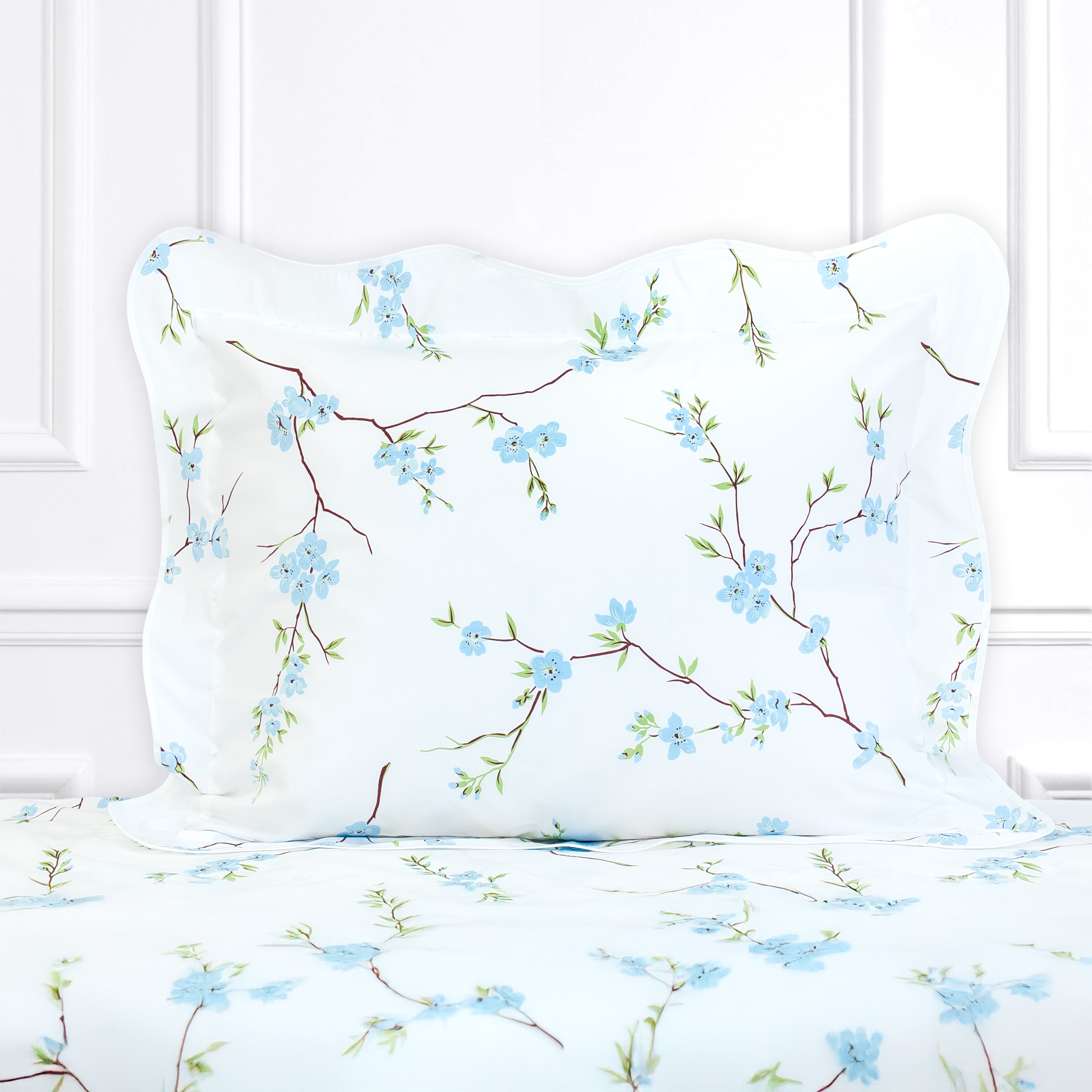 Blue floral with green leaves and brown stems, Fleurs de Pecher print shown on a Standard Sham with a wavy white bias, on top of matching bed sheet with a white panel background.
