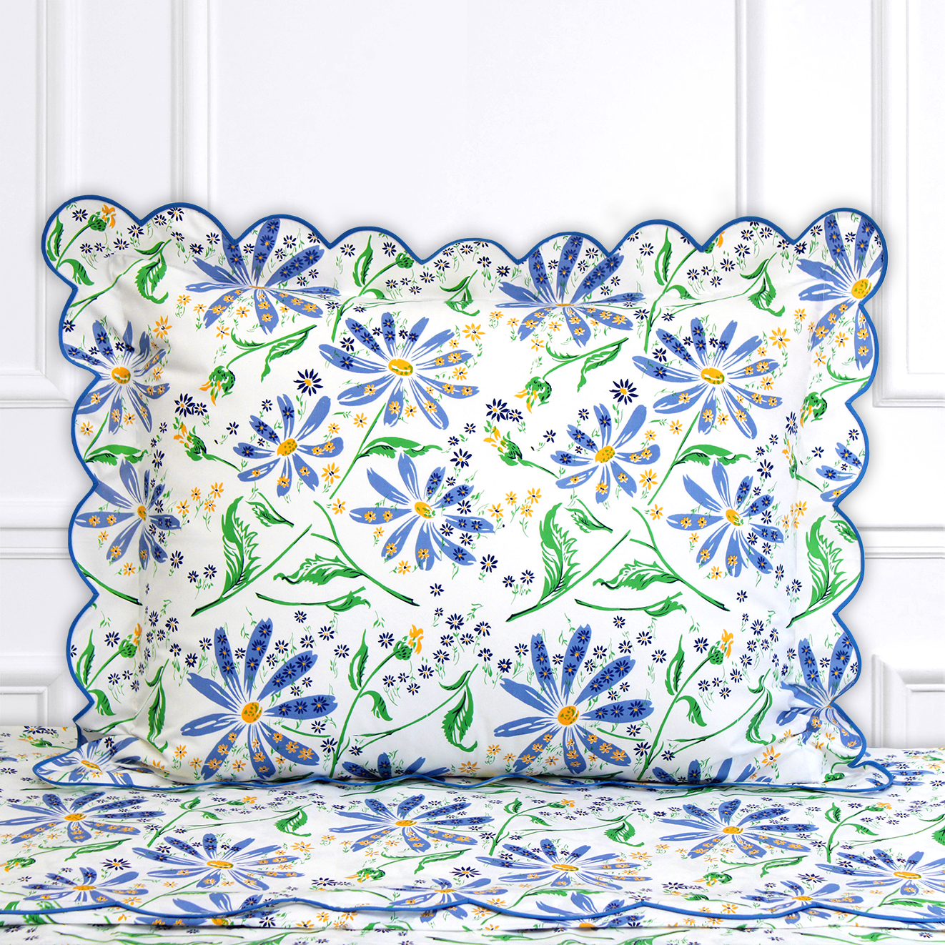 Blue florals with mini yellow florals and green leaves/stems, Marguerites print, shown on a Standard Sham with a scallop blue bias, on top of matching bed sheet with a white panel background.