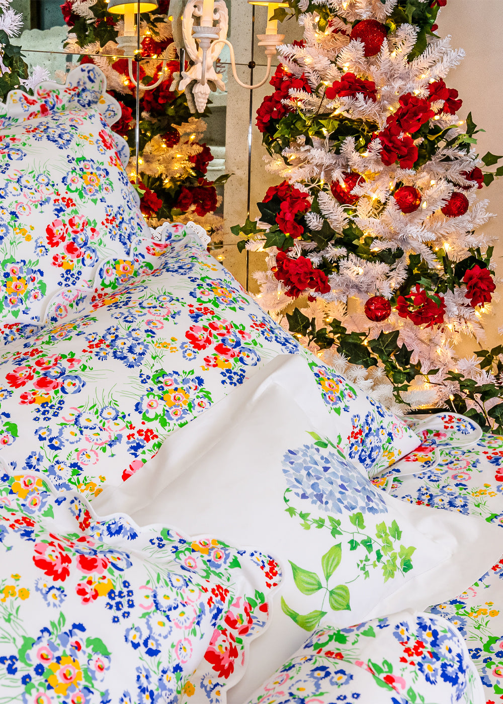 New York Mille Fleurs Multi bed linens styled with an Emb Mariage accent pillow, featuring a colorful allover floral print on crisp white cotton with signature scalloped edges in a festive bedroom setting.