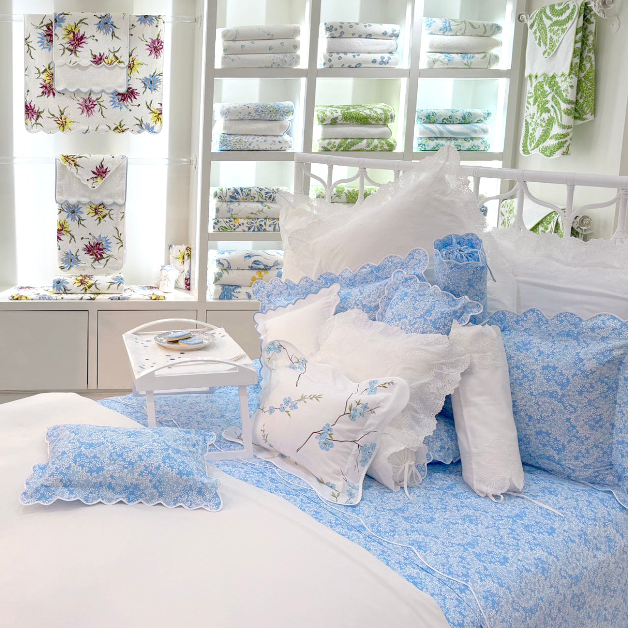 A neatly made bed with blue and white patterned pillows and bedding, next to a small tray with tea and cookies, in front of shelves displaying folded linens.