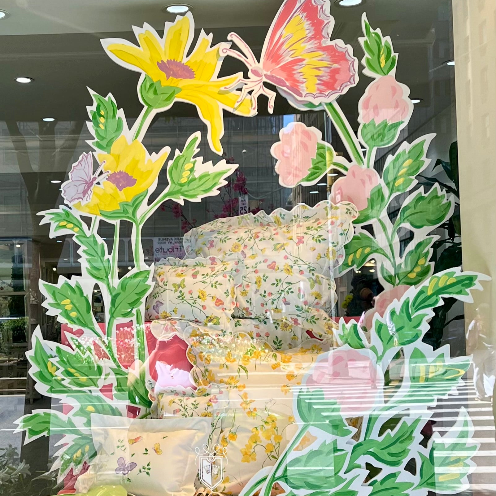 Boutique window display featuring stacked florals printed bedding linens in soft spring colors, framed by large illustrated flowers and butterflies (by Goeff Howell). A folded lime green throw and embroidered shams on the foreground, with the D. Porthault Paris logo visible on the front window.