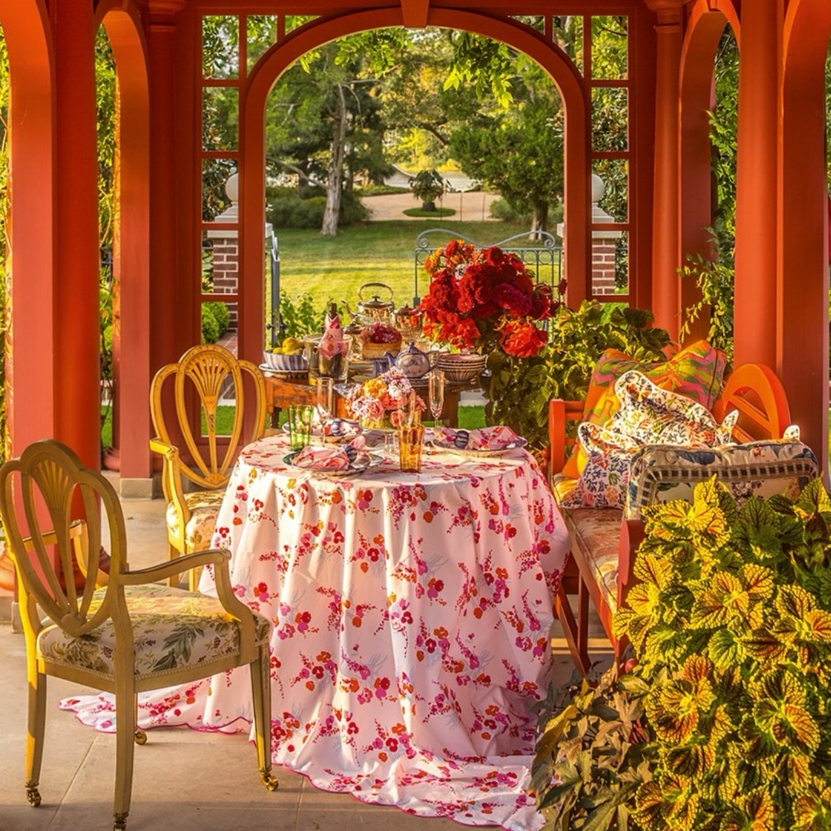 A garden table draped in D. Porthault’s Demoiselles Orange/Pink tablecloth, set with flowers, glassware, and tea service under a terracotta pavilion surrounded by greenery.