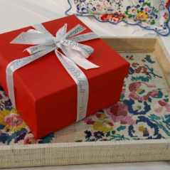 Red gift box tied with a branded white ribbon, styled on a floral laminated tray featuring a colorful allover pattern, with a coordinating scalloped textile in the background.