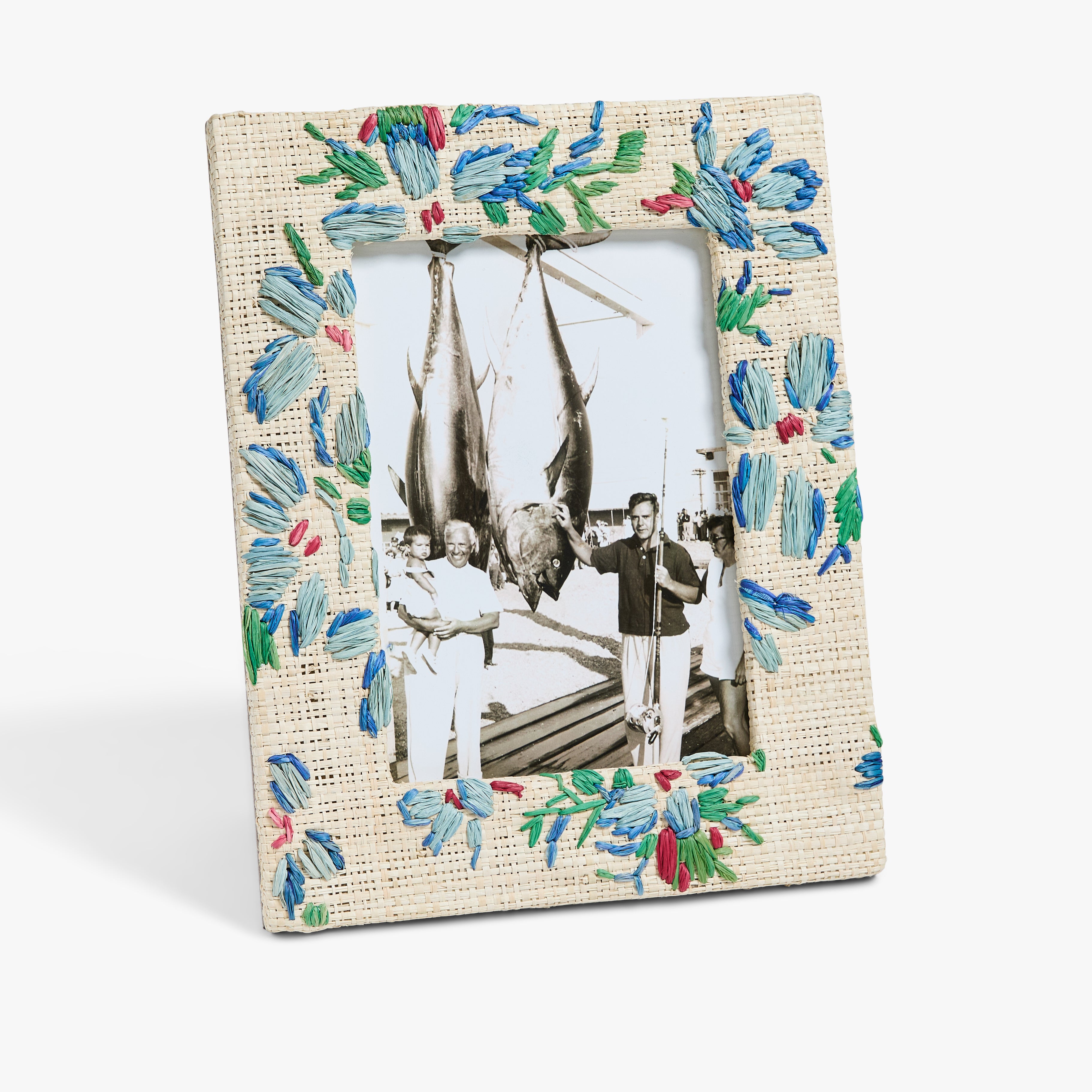 A black-and-white photo of two men standing beside large hanging fish, displayed in a blue and green floral-patterned picture frame.